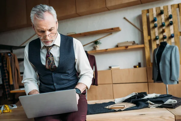 Handsome mature tailor working with laptop while sitting on table at sewing workshop — Stock Photo