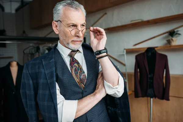 Sastre maduro con confianza en ropa elegante mirando hacia otro lado en el taller de costura - foto de stock