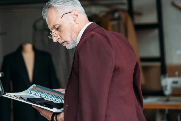 Концентрированный зрелый портной с каталогом типов ткани и пиджака на плечах в швейной мастерской — стоковое фото