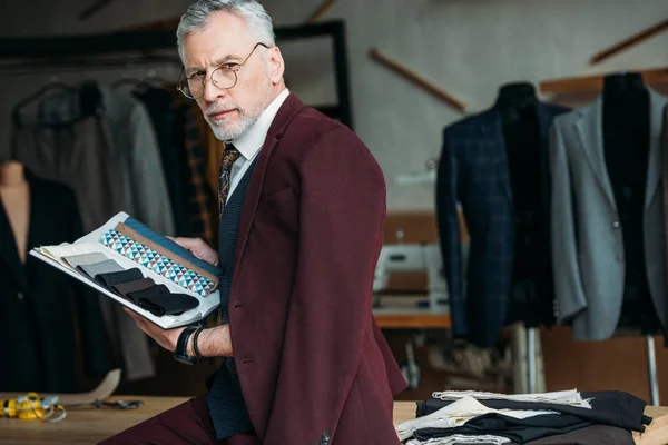 Stylish mature tailor with catalogue of cloth types and jacket on shoulders sitting on table at sewing workshop — Stock Photo