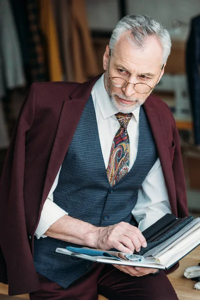 Stylish mature tailor with catalogue of cloth types sitting on table at sewing workshop — Stock Photo