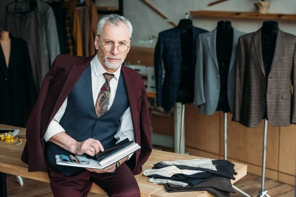 Schöne reife Schneiderin mit Katalog der Stofftypen sitzt auf dem Tisch in der Nähwerkstatt — Stockfoto