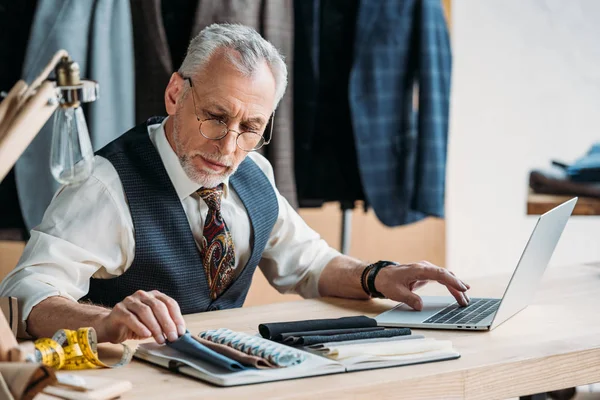 Handsome mature tailor working with laptop and cloth samples at sewing workshop — Stock Photo