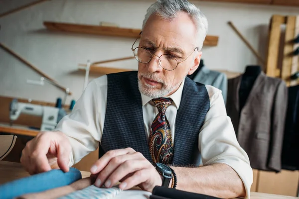 Handsome mature tailor looking at cloth samples at sewing workshop — Stock Photo