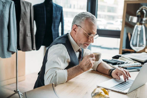 Focused mature tailor working with laptop and drinking coffee at sewing workshop — Stock Photo