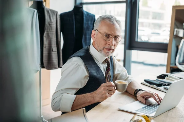 Handsome mature tailor working with laptop and drinking coffee at sewing workshop — Stock Photo
