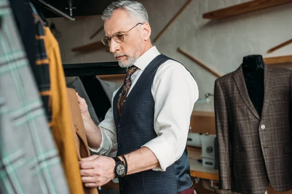 Красивый зрелый портной осматривает куртки в швейной мастерской — стоковое фото