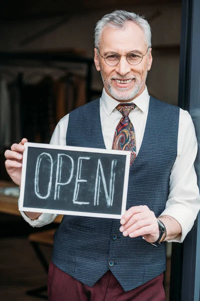 Smiling mature tailor holding chalkboard with open sign at sewing workshop — Stock Photo