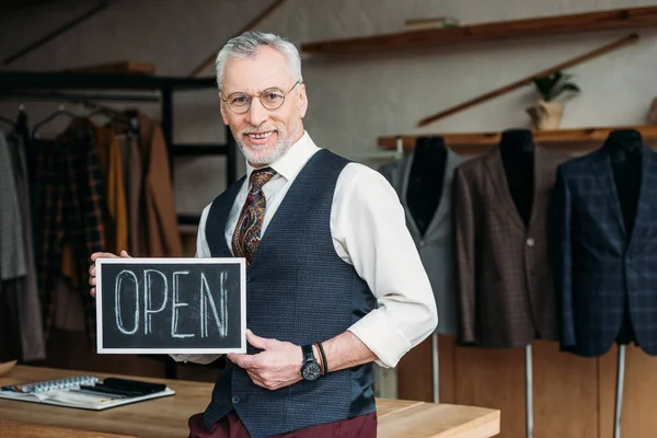 Handsome mature tailor holding chalkboard with open sign at sewing workshop — Stock Photo