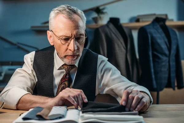 Beau tailleur mature travaillant avec des échantillons de tissu à l'atelier de couture en fin de soirée — Photo de stock