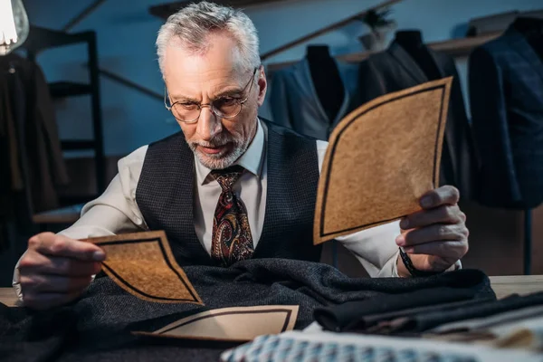 Handsome mature tailor working with cut patterns at sewing workshop in late evening — Stock Photo