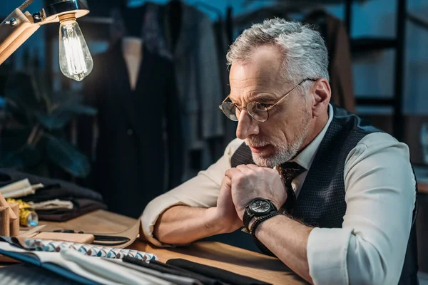 Confident mature tailor looking at cloth samples at sewing workshop in late evening — Stock Photo