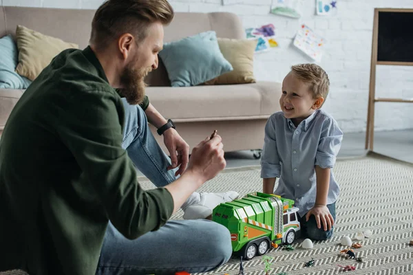 Pai e pequeno filho sorridente brincando com carros de brinquedo juntos no chão em casa — Fotografia de Stock