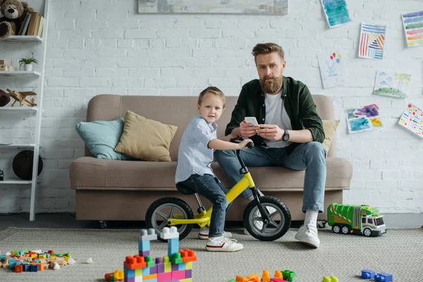 Little kid on balance bicycle with bearded father with smartphone sitting on sofa at home — Stock Photo