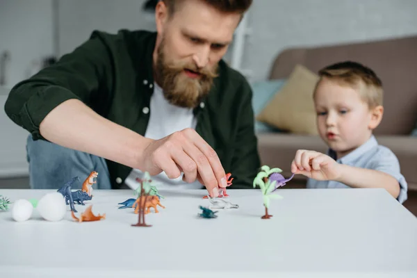Padre y lindo hijo pequeño jugando con varios dinosaurios de juguete juntos en casa - foto de stock