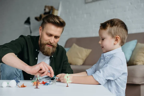 Padre y lindo hijo pequeño jugando con varios dinosaurios de juguete juntos en casa - foto de stock