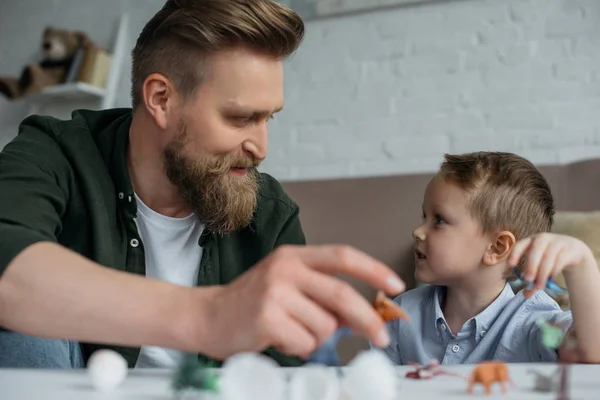 Padre y lindo hijo pequeño jugando con varios dinosaurios de juguete juntos en casa - foto de stock