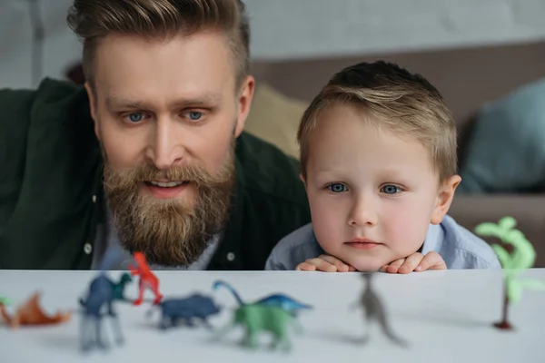 Selective focus of father and cute little son looking at arranged toy dinosaurs on tabletop together at home — Stock Photo