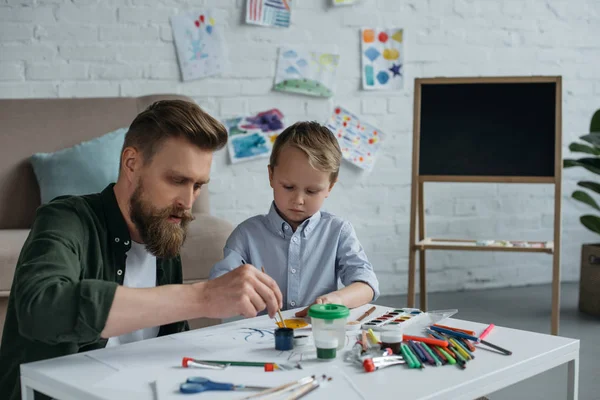 Fokussierter Vater und niedlicher kleiner Sohn mit Farben und Pinseln, die zu Hause gemeinsam Bilder malen — Stockfoto