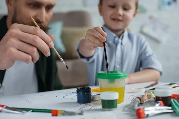 Selektiver Fokus von Vater und niedlichem kleinen Sohn mit Farben und Pinseln, die Bilder zu Hause zusammenzeichnen — Stockfoto