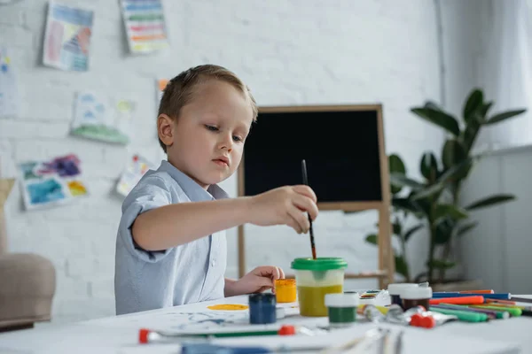 Focused little boy with brush and paintings drawing picture alone at home — Stock Photo