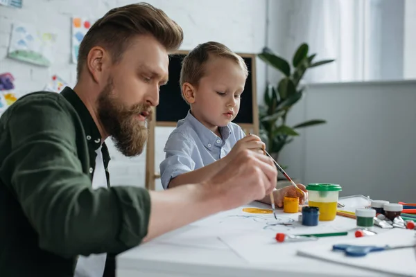 Vista laterale del padre e del piccolo figlio carino con vernici e pennelli che disegnano insieme i quadri a casa — Foto stock