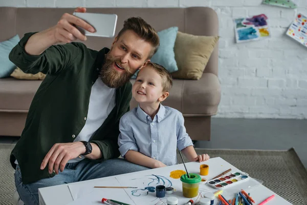 Padre e figlio sorridenti che si fanno selfie sullo smartphone insieme a casa — Foto stock