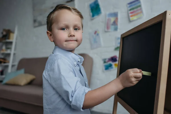 Vista lateral do menino com pedaço de giz olhando para a câmera ao desenhar imagem no quadro em casa — Fotografia de Stock