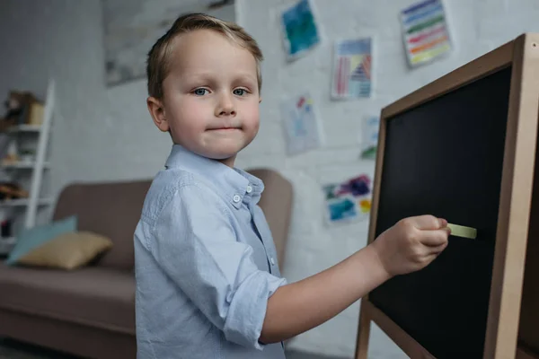 Seitenansicht eines kleinen Jungen mit einem Stück Kreide, der in die Kamera schaut, während er zu Hause ein Bild auf der Tafel zeichnet — Stockfoto