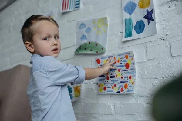 Kleines Kind blickt in Kamera, während es zu Hause kindliche Bilder an die Wand hängt — Stockfoto