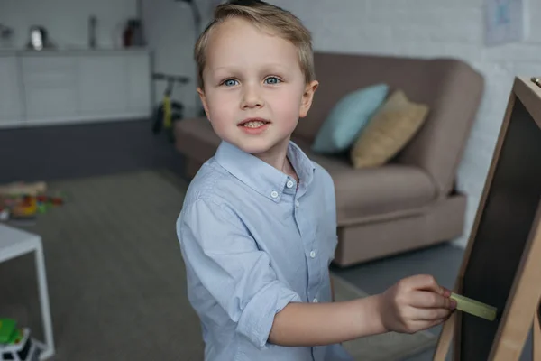 Vista lateral del niño pequeño con un pedazo de tiza mirando a la cámara mientras dibuja la imagen en la pizarra en casa - foto de stock