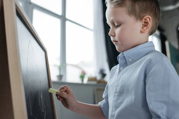 Side view of little boy with piece of chalk drawing picture on blackboard at home — Stock Photo