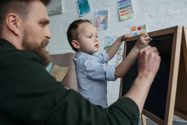 Vista lateral del niño y el padre con trozos de tiza dibujo en pizarra en casa - foto de stock