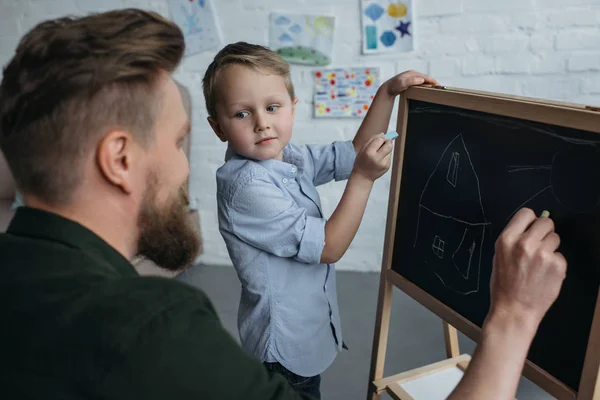 Vista lateral del niño y el padre con trozos de tiza dibujo en pizarra en casa - foto de stock