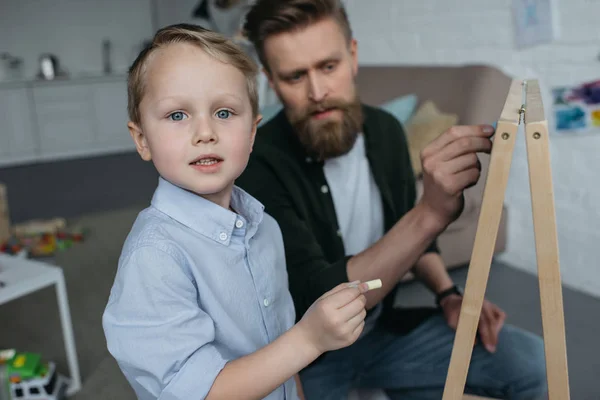Selective focus of little boy and father with pieces of chalk drawing picture on blackboard at home — Stock Photo