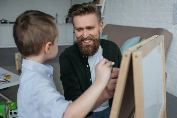 Niño y padre sonriente con trozos de tiza dibujando en pizarra en casa - foto de stock