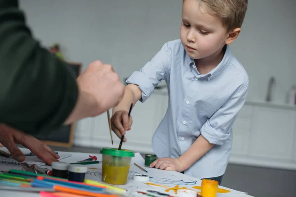 Cropped shot of little boy with brush and paints drawing picture together with father at home — Stock Photo