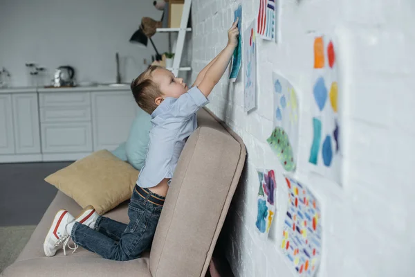 Vue latérale du petit enfant accrochant des photos enfantines sur le mur à la maison — Photo de stock