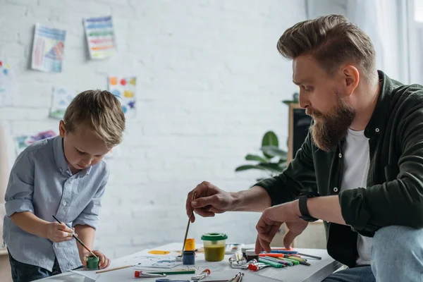 Kleiner Junge mit Pinsel und Farben zeichnet Bild zusammen mit Vater zu Hause — Stockfoto