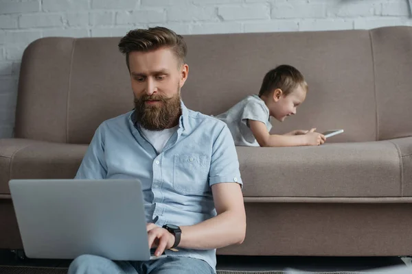 Bearded man using laptop while son with tablet lying on sofa at home — Stock Photo