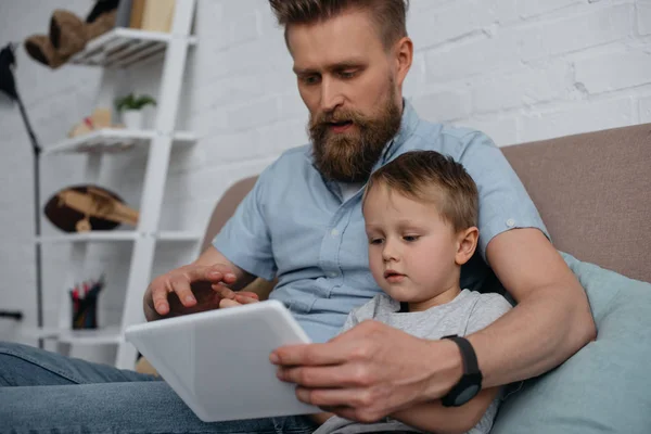 Pai focado e pequeno filho usando tablet juntos no sofá em casa — Fotografia de Stock