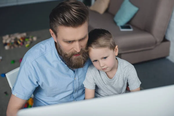Отец и маленький сын смотрит на экран компьютера во время использования компьютера дома — стоковое фото