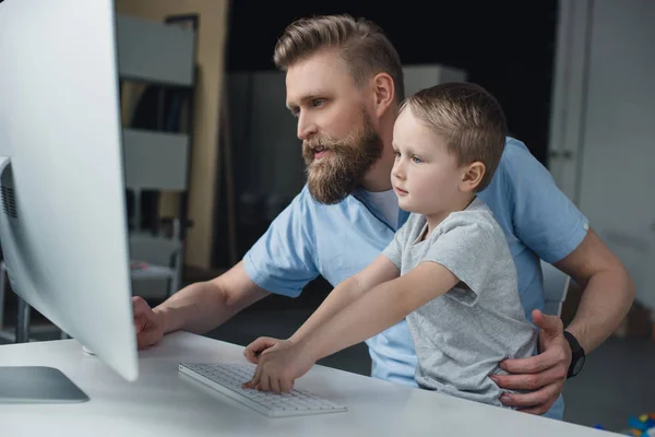 Padre e hijo pequeño mirando la pantalla de la computadora mientras usa la computadora en casa - foto de stock