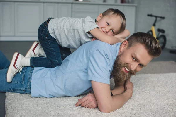 Side view of smiling son lying on fathers back at home — Stock Photo