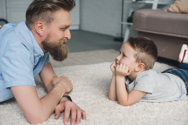 Vue latérale du père barbu et du fils souriant se regardant à la maison — Photo de stock