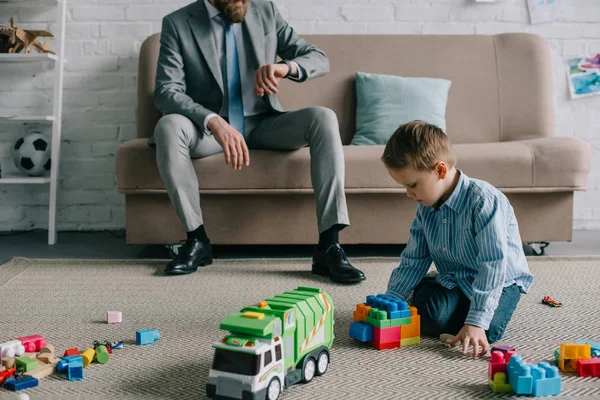 Teilansicht eines Geschäftsmannes, der die Zeit kontrolliert, während Sohn zu Hause mit Spielzeug auf dem Boden spielt, Arbeits- und Life-Balance-Konzept — Stockfoto