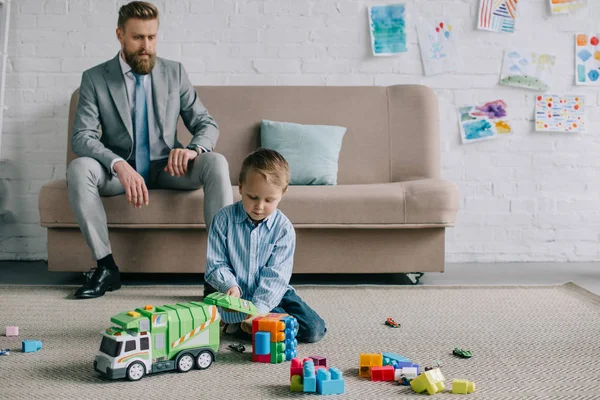 Geschäftsmann im Anzug mit Blick auf den kleinen Sohn, der zu Hause mit bunten Bauklötzen spielt, Arbeits- und Life-Balance-Konzept — Stockfoto