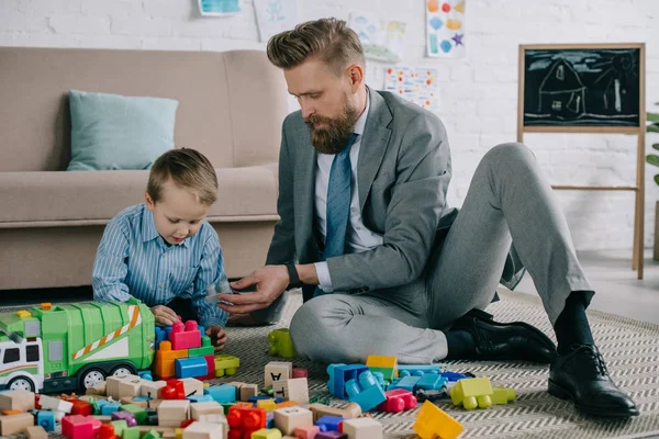 Businessman in suit and little son playing with colorful blocks together on floor at home, work and life balance concept — Stock Photo