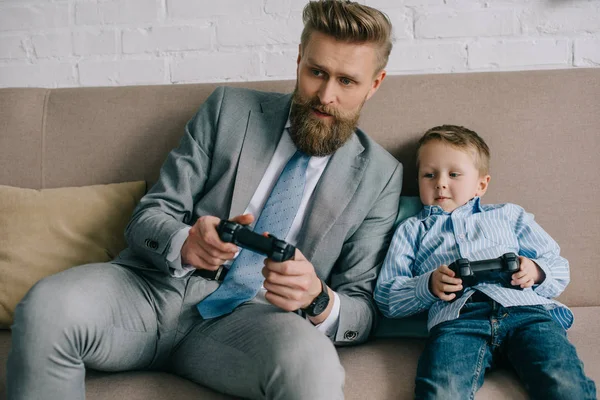 Hombre de negocios e hijo pequeño jugando videojuegos juntos en el hogar, el trabajo y el equilibrio de la vida concepto - foto de stock