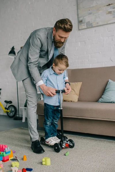 Hombre de negocios en traje e hijo pequeño en scooter en el hogar, el trabajo y el equilibrio de la vida concepto - foto de stock